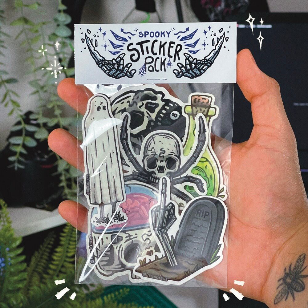Mano sosteniendo un paquete de pegatinas con dibujos de temática terrorífica y una cabecera que dice «SPOOKY STICKER PACK», sobre un fondo de plantas.