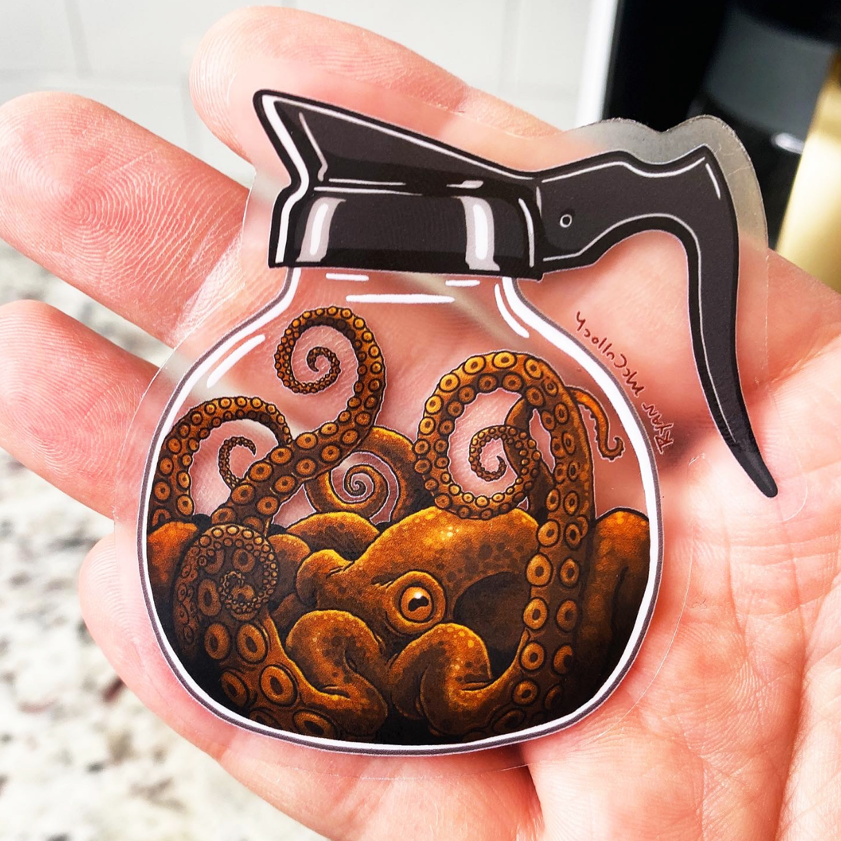 Die-cut transparent sticker showing an octopus in a coffee pot, held up by a hand in front of a stone floor