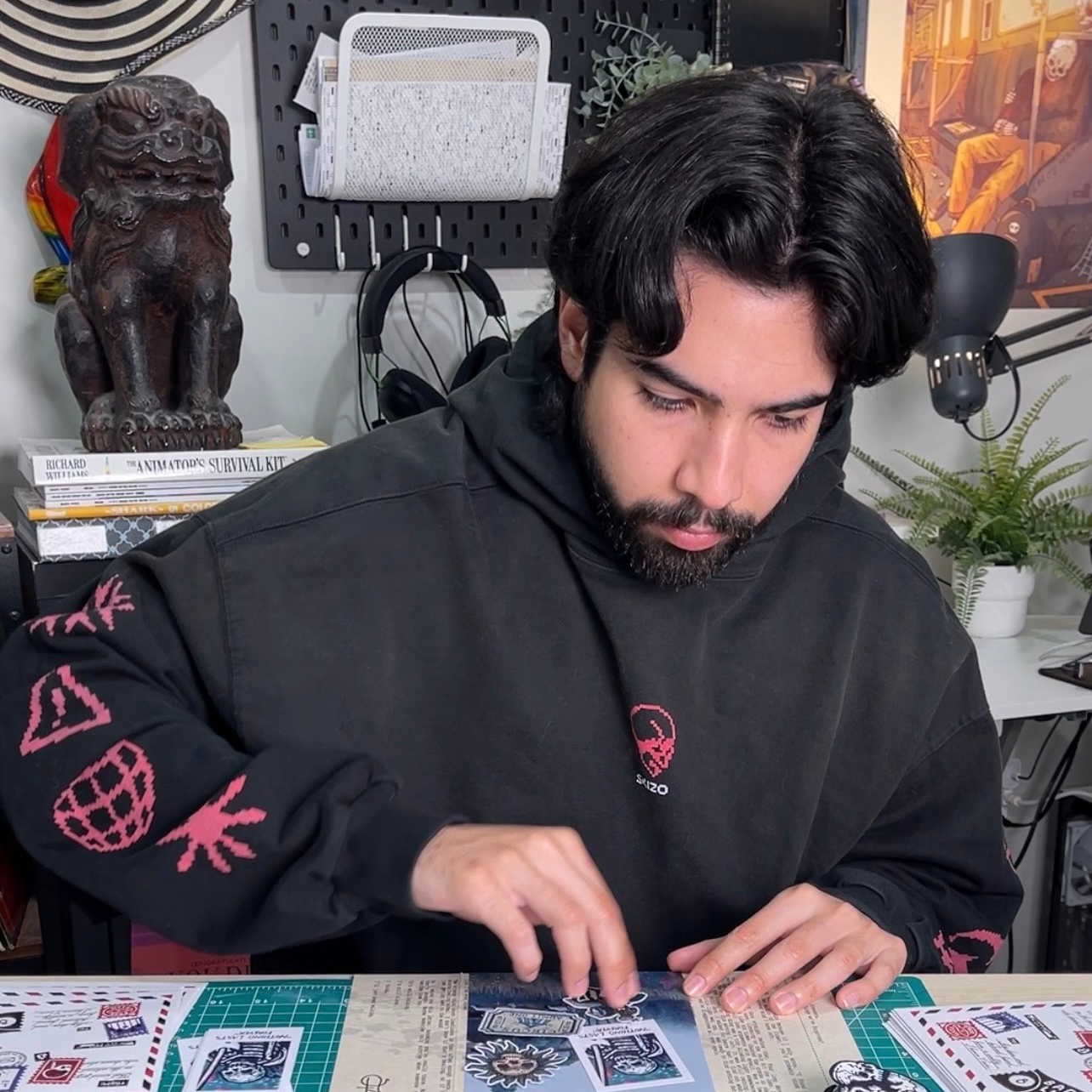 Un hombre con sudadera negra con capucha sentado en su estudio, colocando pegatinas sobre la mesa, con esculturas y obras de arte al fondo.