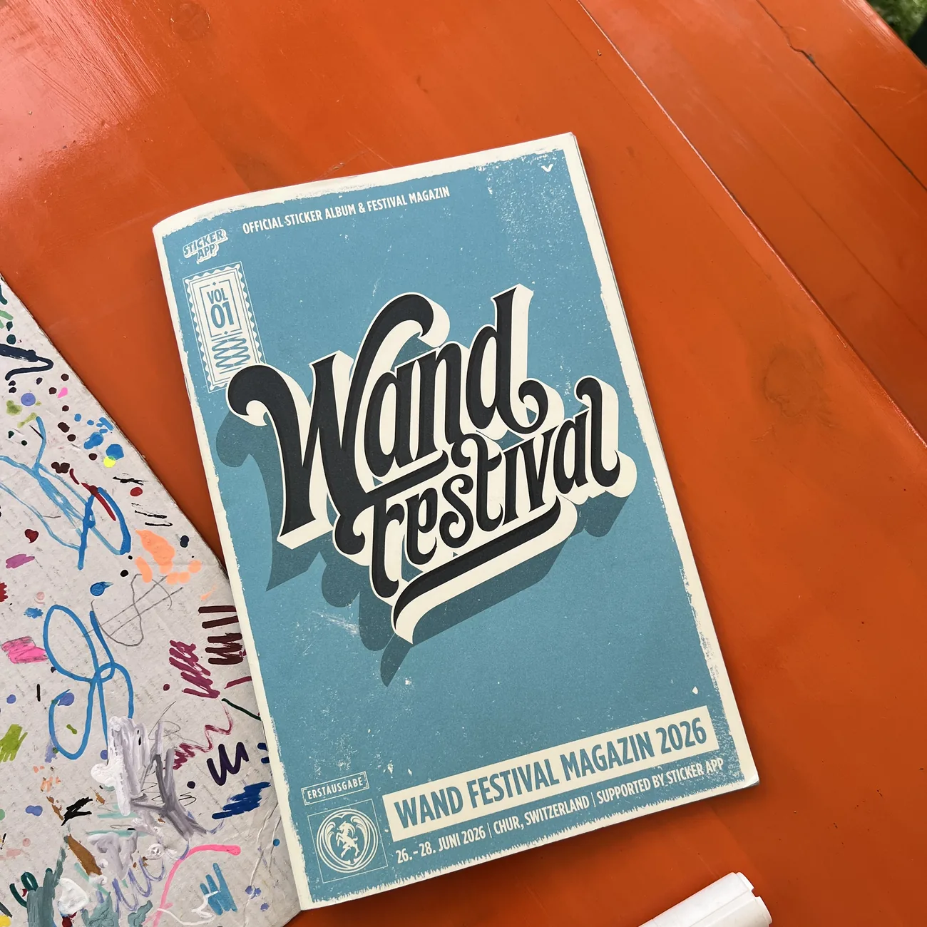 Álbum oficial de pegatinas y revista del Wand Festival 2026, con el apoyo de StickerApp, junto a un rotulador de pintura sobre una mesa naranja