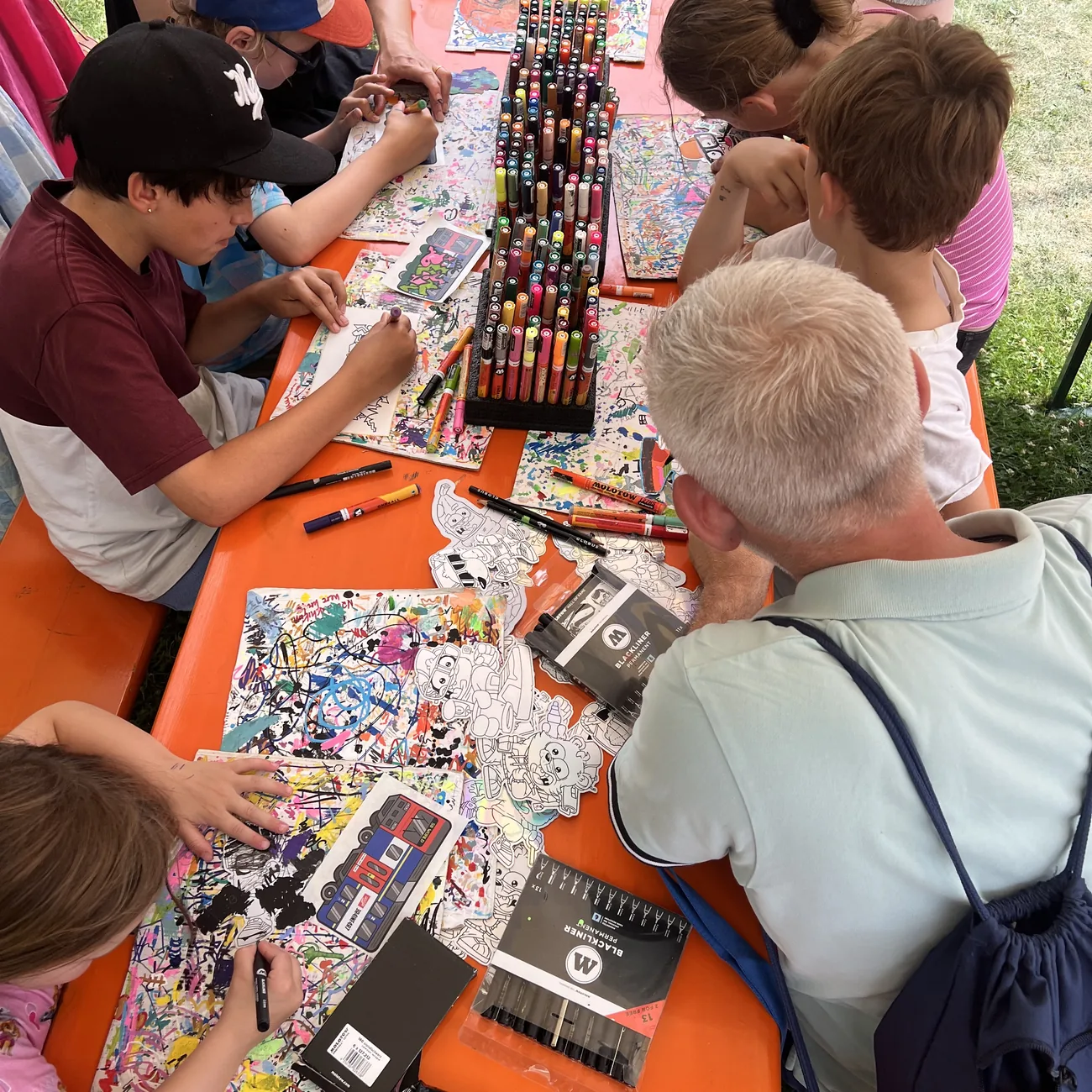 Niños coloreando pegatinas troqueladas de Brainfart alrededor de un soporte de rotuladores de pintura en un taller de festival al aire libre