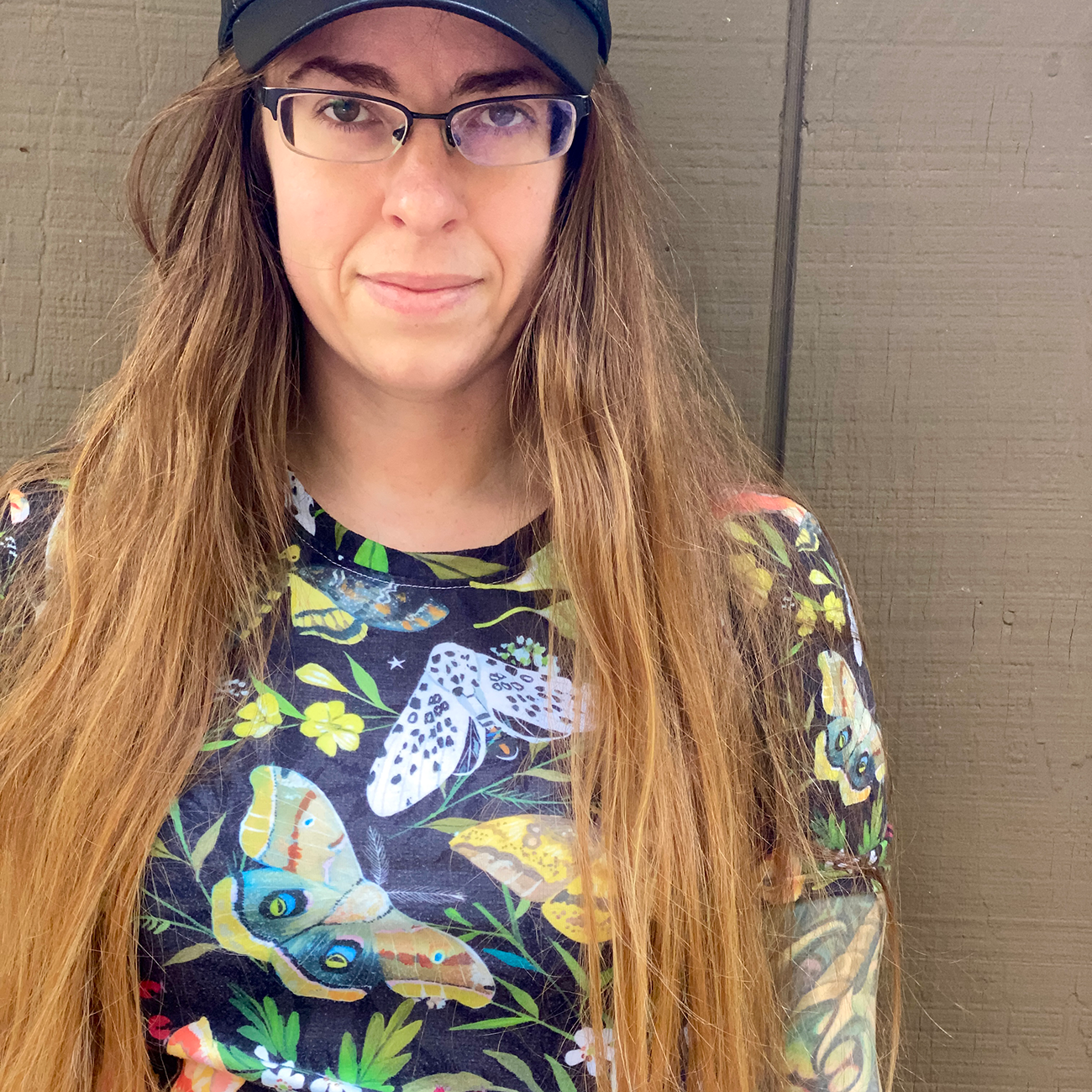 Lana Ansay aka oddarette with long hair and glasses wearing a cap and a colorful shirt with moth designs.