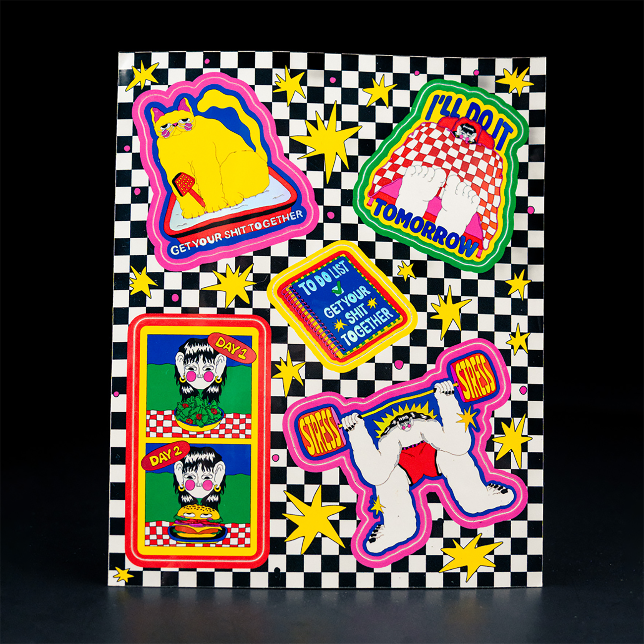 Colorful sticker sheet with bold, playful designs on a black-and-white checkerboard background. Features quirky illustrations including a yellow cat, a gorilla, and surreal characters, surrounded by stars and bright graphic elements.