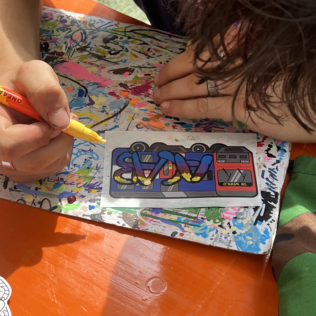 Una mano colorea una etiqueta del tren S8 World con un rotulador de pintura amarillo en el taller de Brainfart x StickerApp