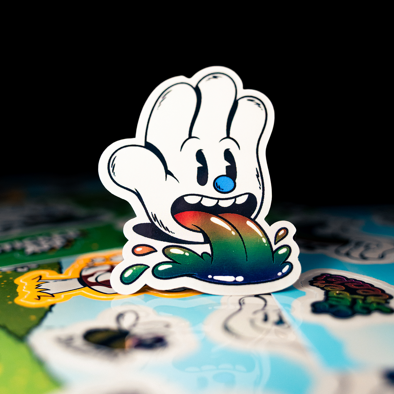 Cartoon sticker of a hand-shaped character with a blue nose, open mouth, and rainbow-colored tongue, on a surface with various other stickers.