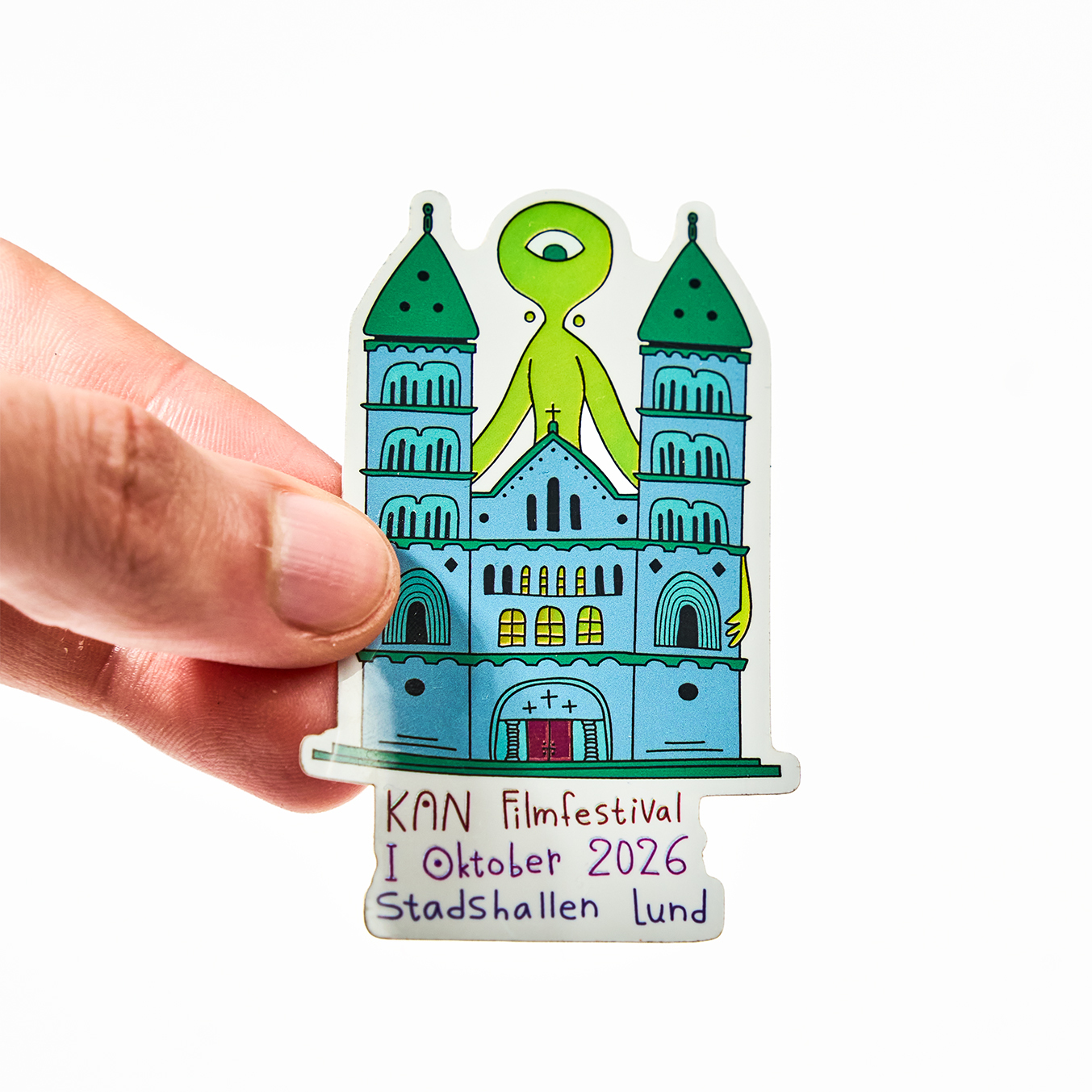 Mano sosteniendo una pegatina troquelada para el KAN Filmfestival, octubre de 2026 en Stadshallen Lund, con una catedral y un extraterrestre verde de un solo ojo