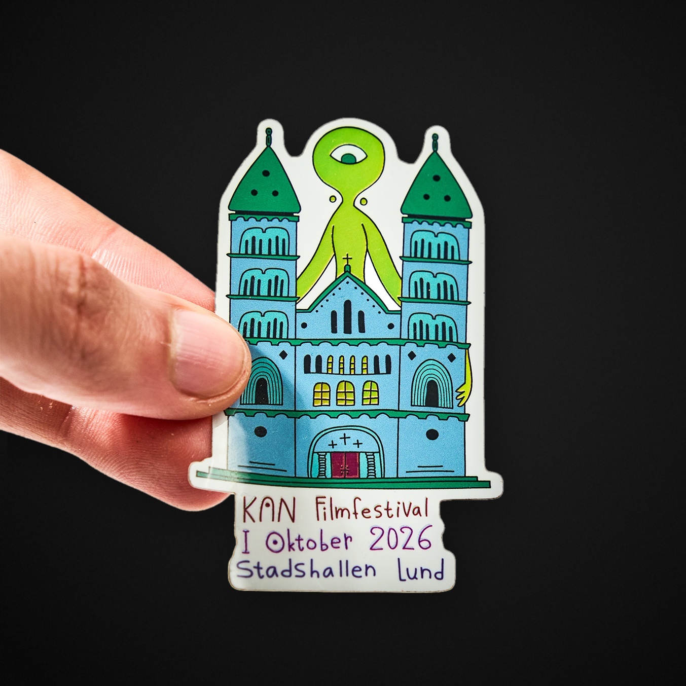 Mano sujetando una pegatina troquelada de una catedral con una criatura verde de un solo ojo y el texto «KAN Filmfestival I Oktober 2026 Stadshallen Lund»