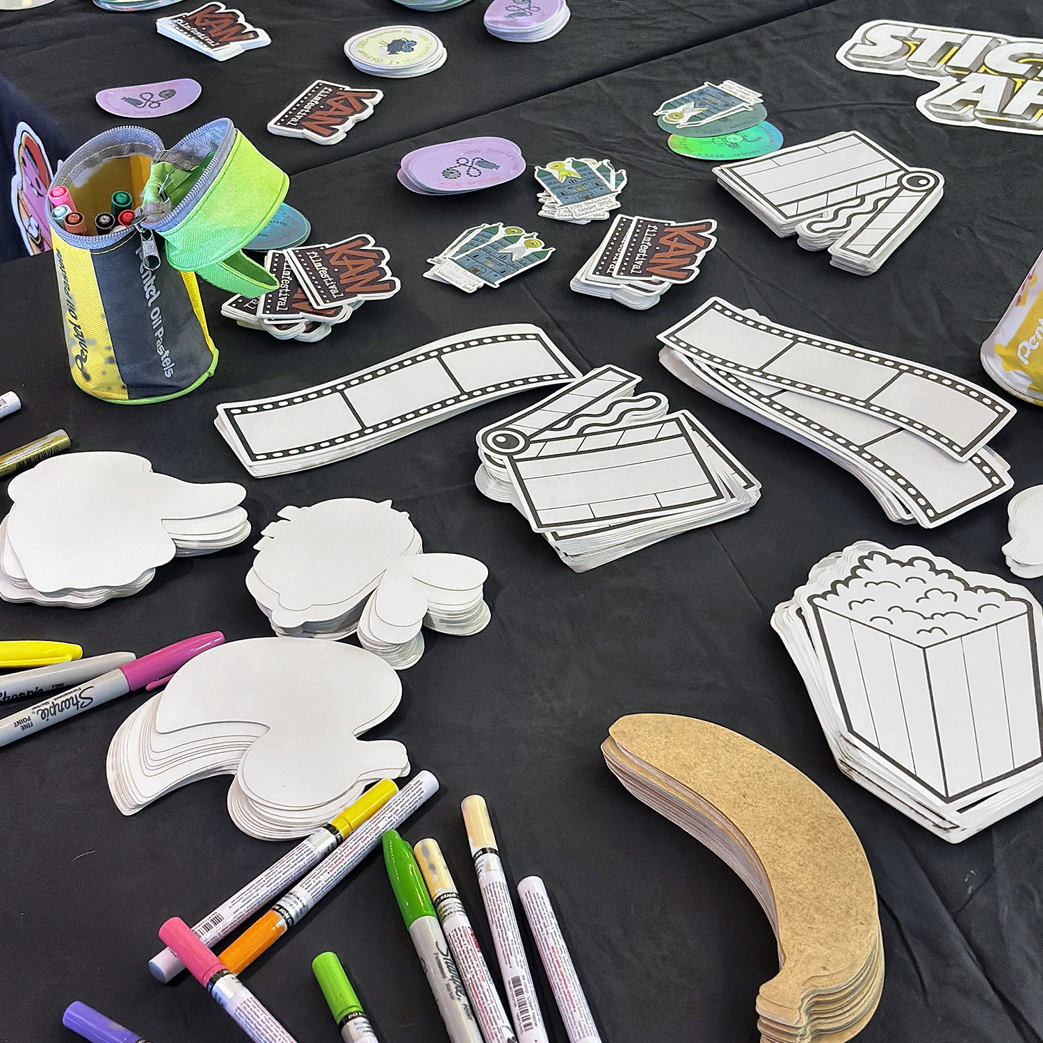 Mesa para crear pegatinas en el KAN Film Festival con pegatinas troqueladas en blanco con forma de palomitas, tiras de película y claquetas, pegatinas de papel kraft y rotuladores de pintura