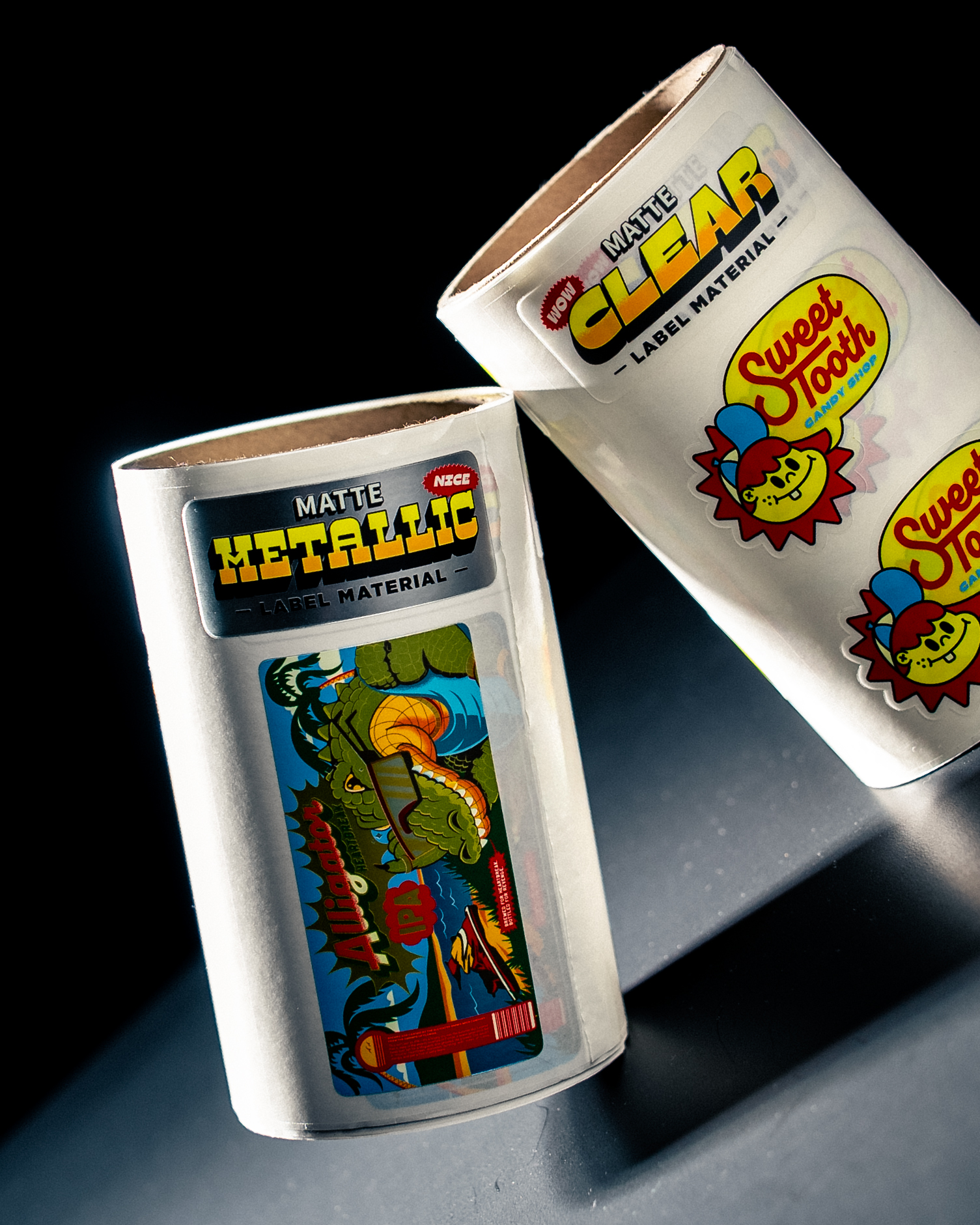 Dos rollos de etiquetas de muestra con los rótulos «Matte Metallic» y «Matte Clear», con una etiqueta de Alligator IPA y etiquetas de Sweet Tooth Candy Shop