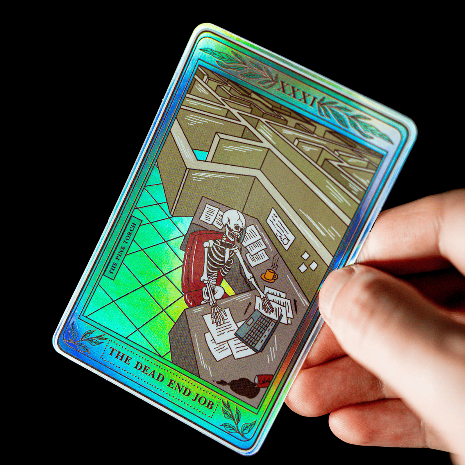 Mano sujetando una pegatina holográfica de carta del tarot titulada "The Dead End Job", con un esqueleto sentado en un escritorio de oficina