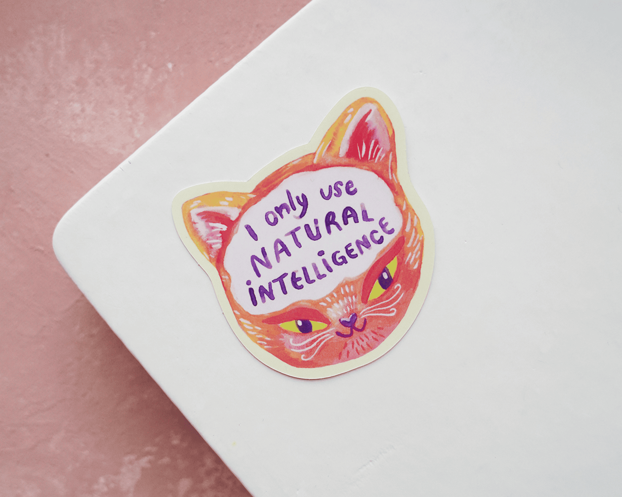 Pegatina troquelada de una cara de gato con el texto "I only use natural intelligence" sobre una libreta blanca