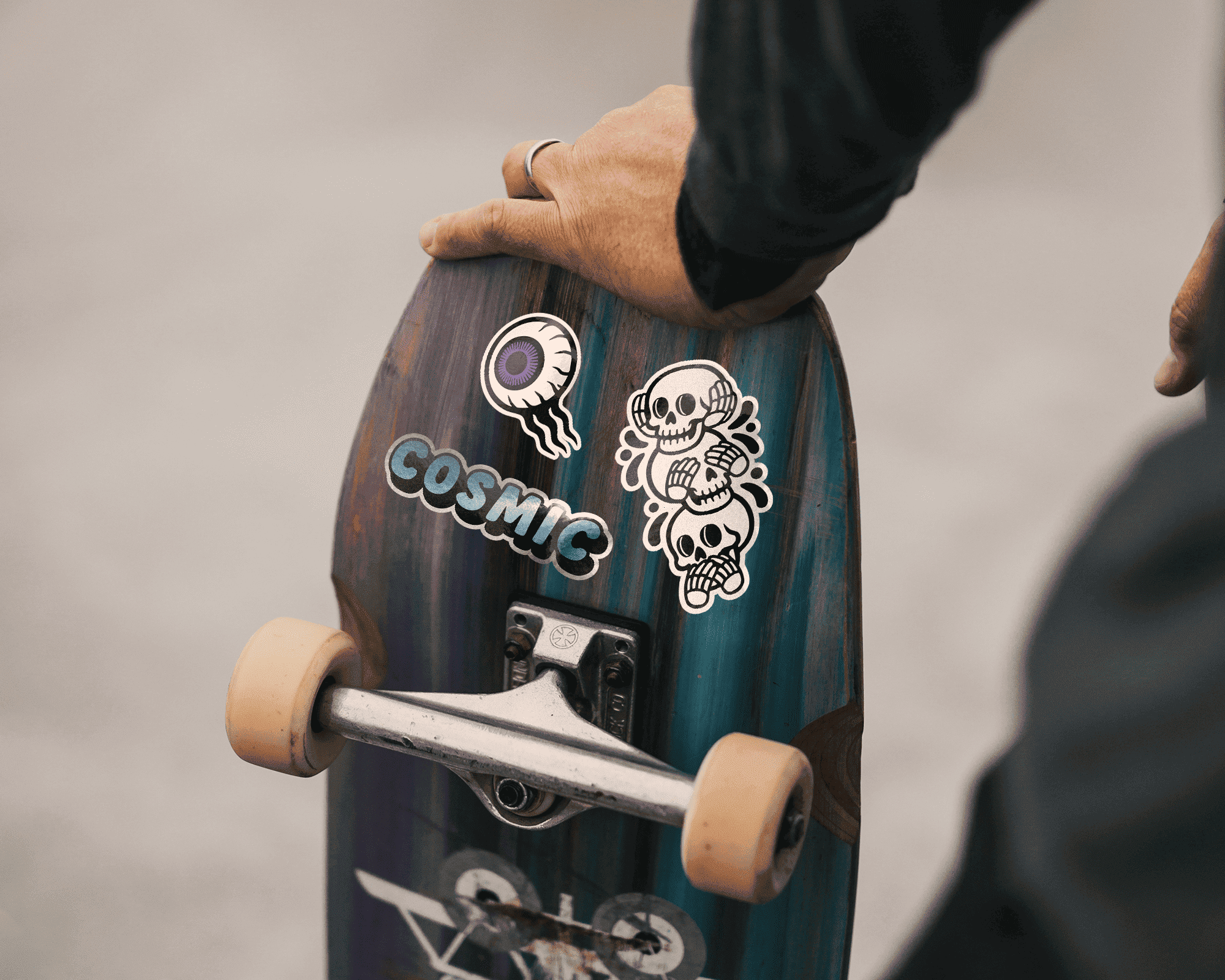 Close-up of a person holding a skateboard with colorful stickers, including an eye and skulls, on a blurred background.