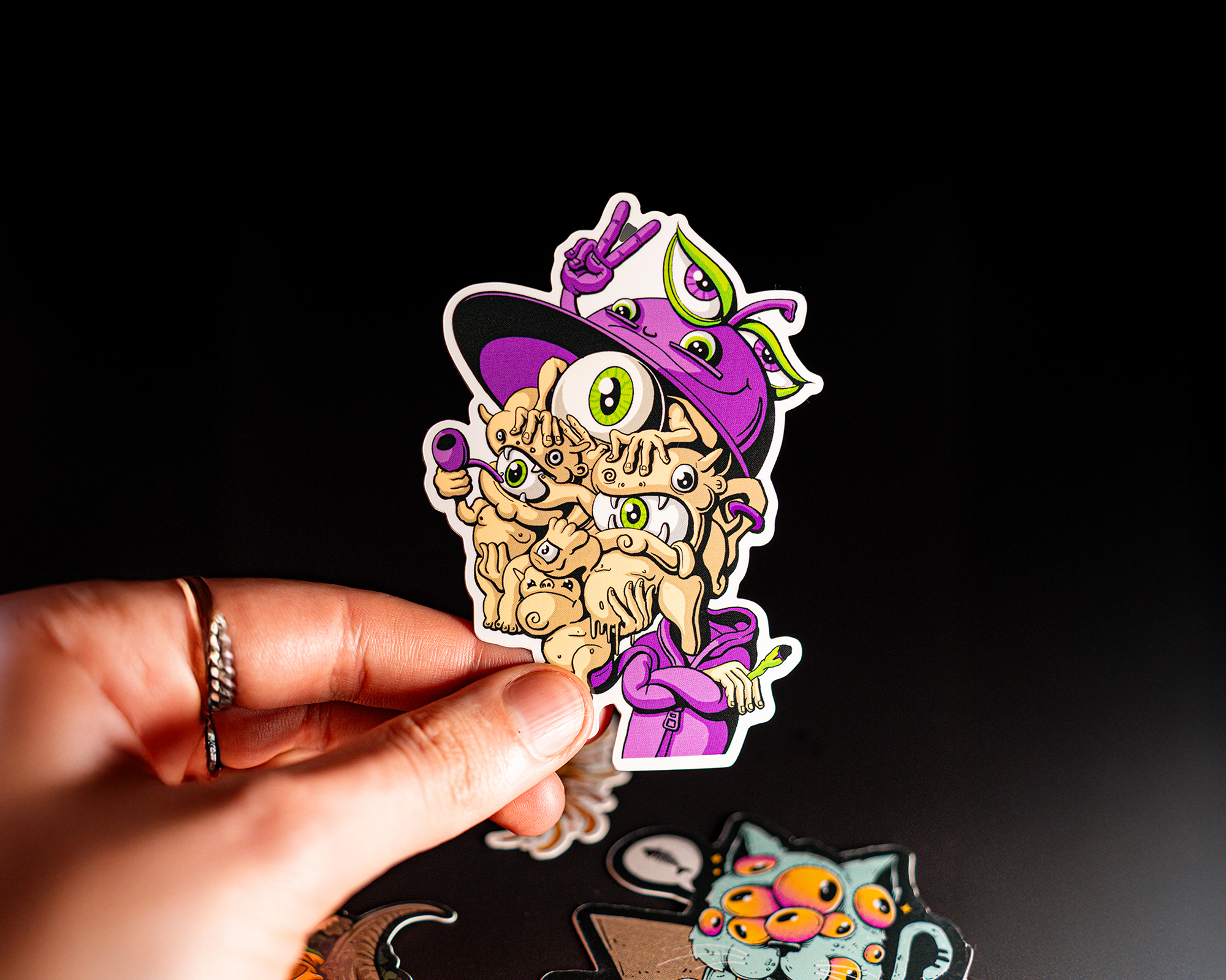 A hand holding a colorful die cut sticker of a cartoon character with multiple eyes, wearing a purple hat, against a dark background.