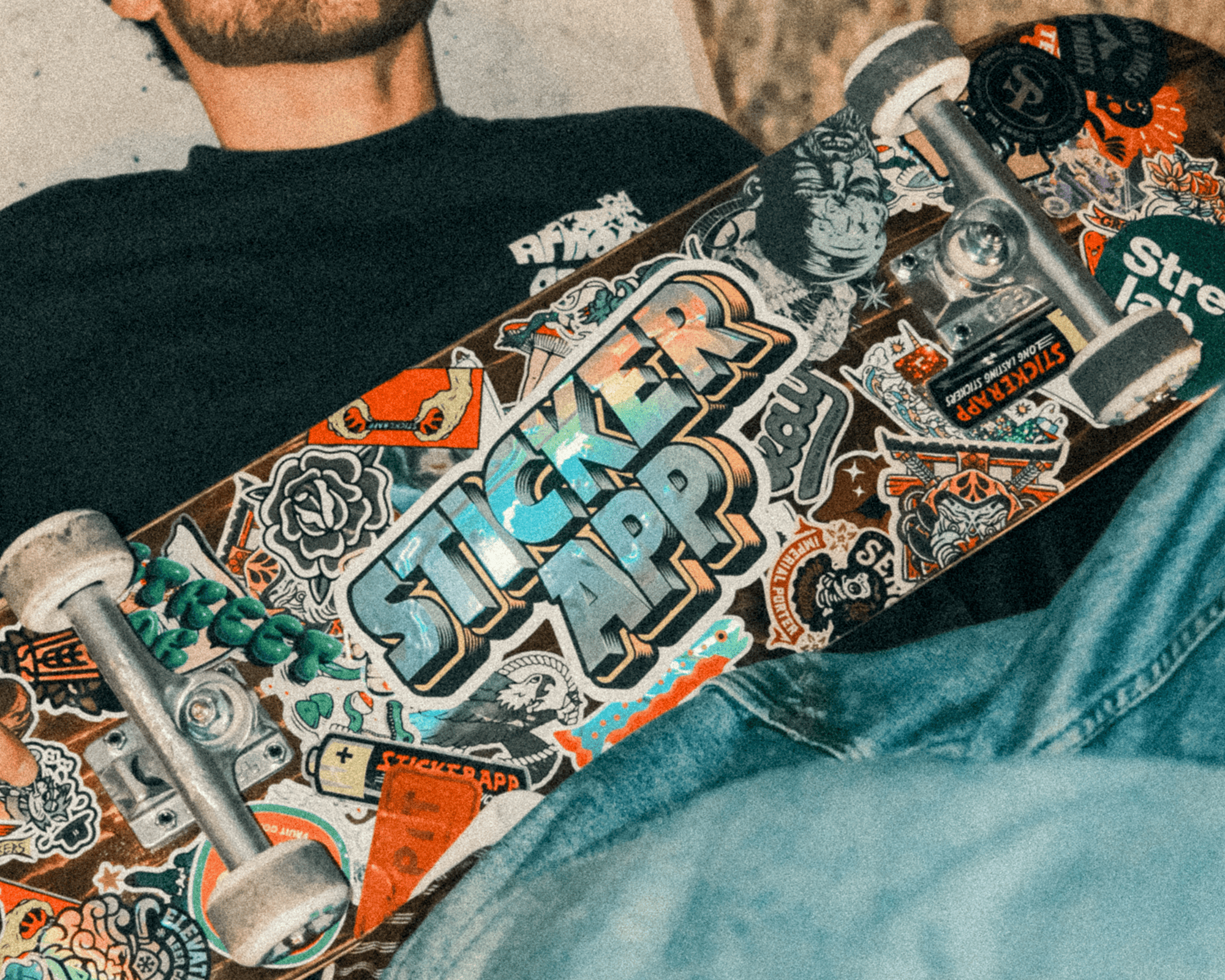 Person holding a skateboard covered in colorful stickers, including a prominent "Sticker App" logo, wearing a dark shirt and jeans.