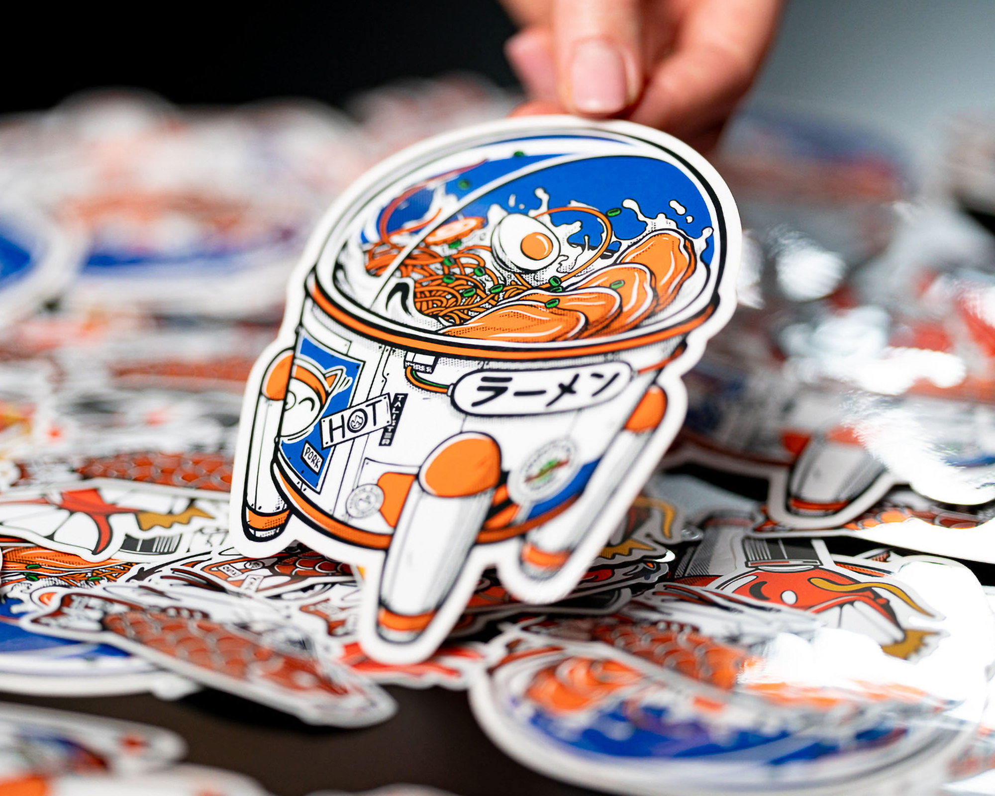 A hand holds a colorful vinyl sticker of an astronaut with a ramen bowl as a helmet, surrounded by similar stickers.