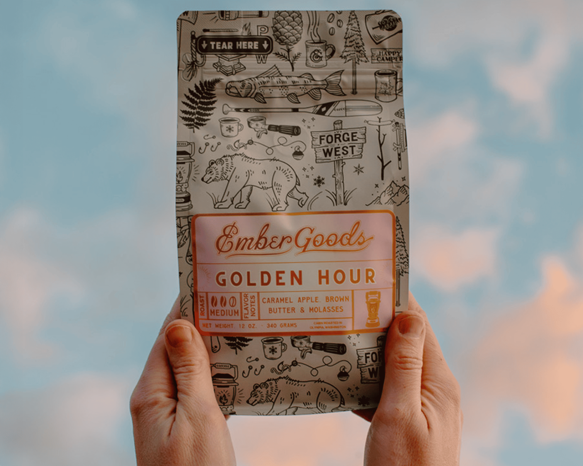 Manos sosteniendo una bolsa de café Golden Hour de Ember Goods con una etiqueta metalizada y el cielo de fondo