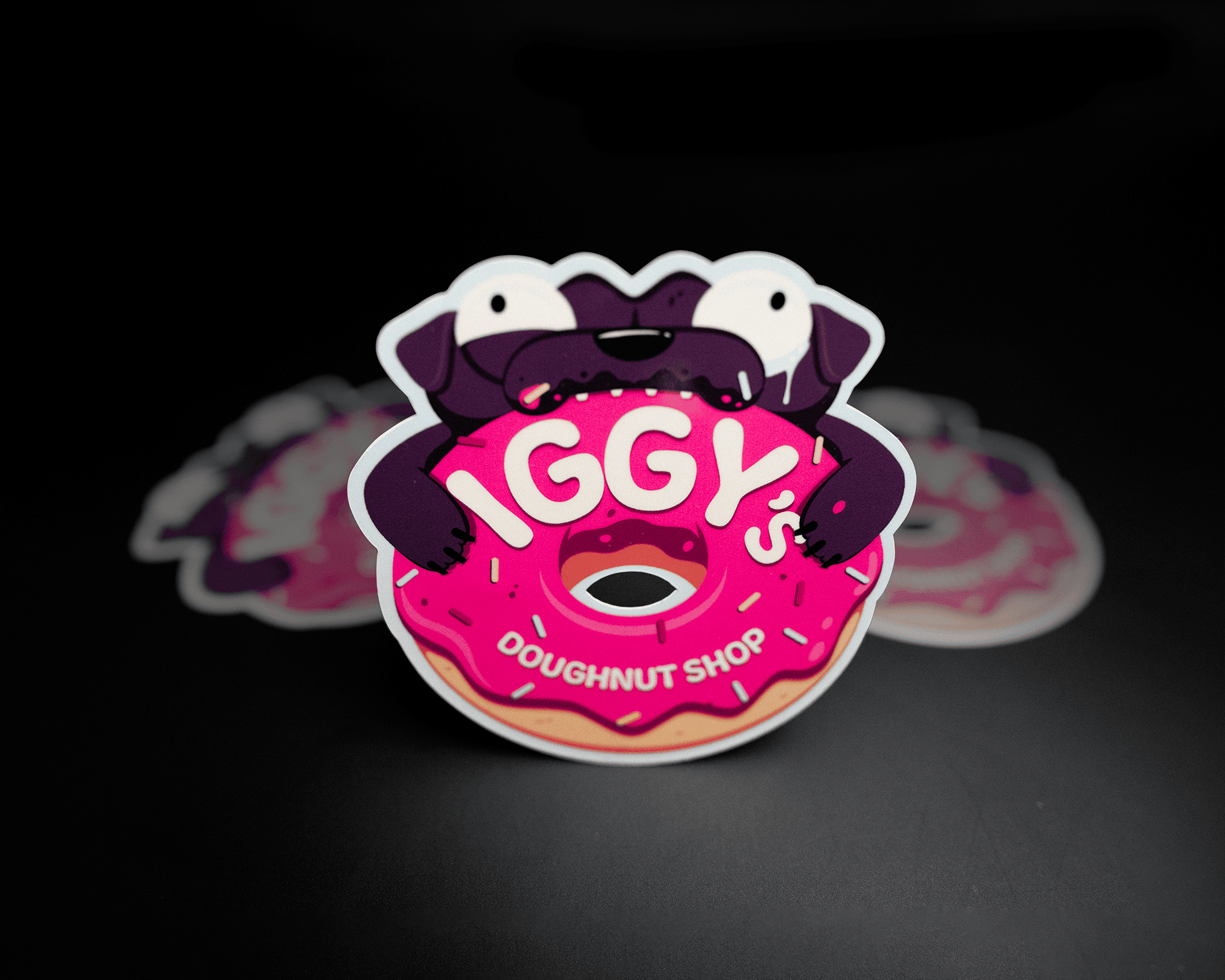A die-cut sticker of a cartoon pug biting a pink frosted donut with "IGGY'S DOUGHNUT SHOP" text on it, set against a dark background.