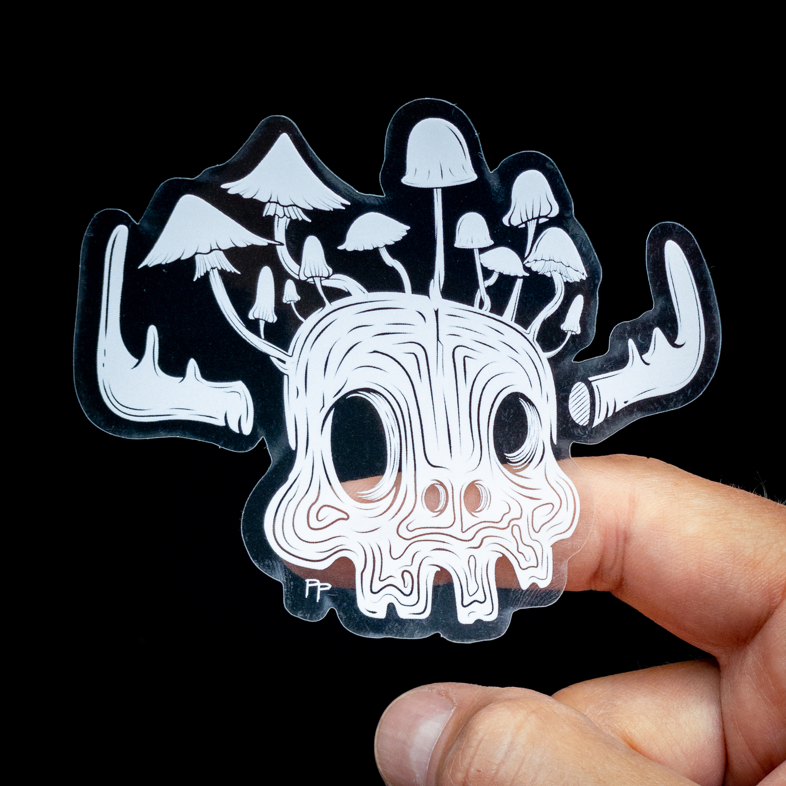 Transparent sticker of a white skull with mushrooms growing from it, held between fingers against a black background.