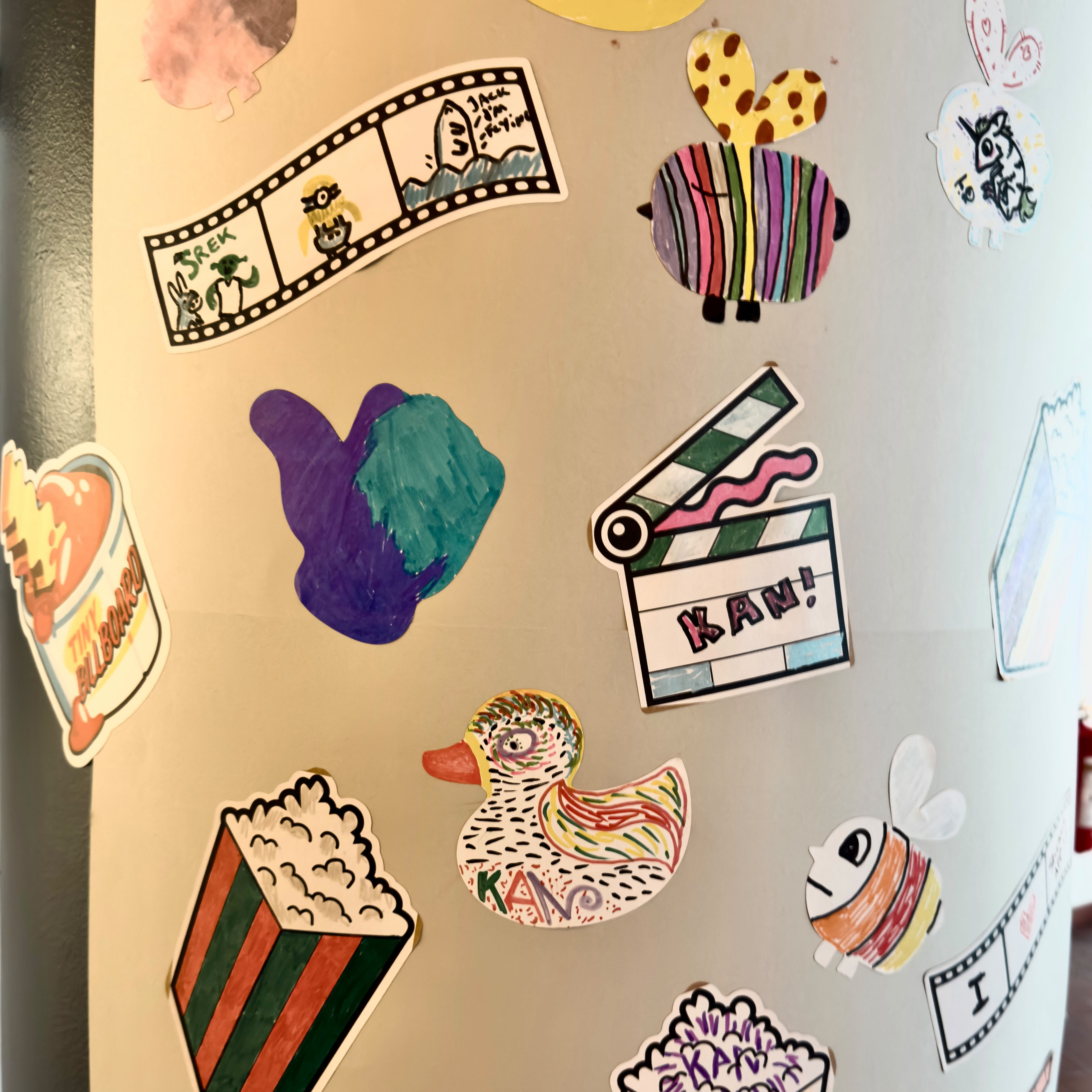 Columna cubierta de pegatinas dibujadas a mano por los visitantes del KAN Film Festival, con palomitas, tiras de película, un pato y una claqueta que dice «KAN!»